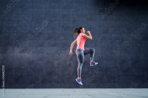 Side view of fit attractive Caucasian woman in sportswear and with ponytail running outdoors. In background is dark wall.