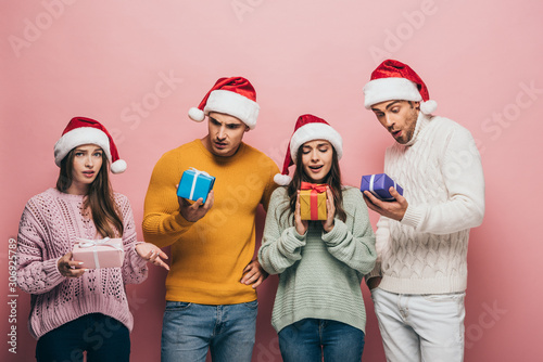surprised friends in santa hats holding christmas presents, isolated on pink