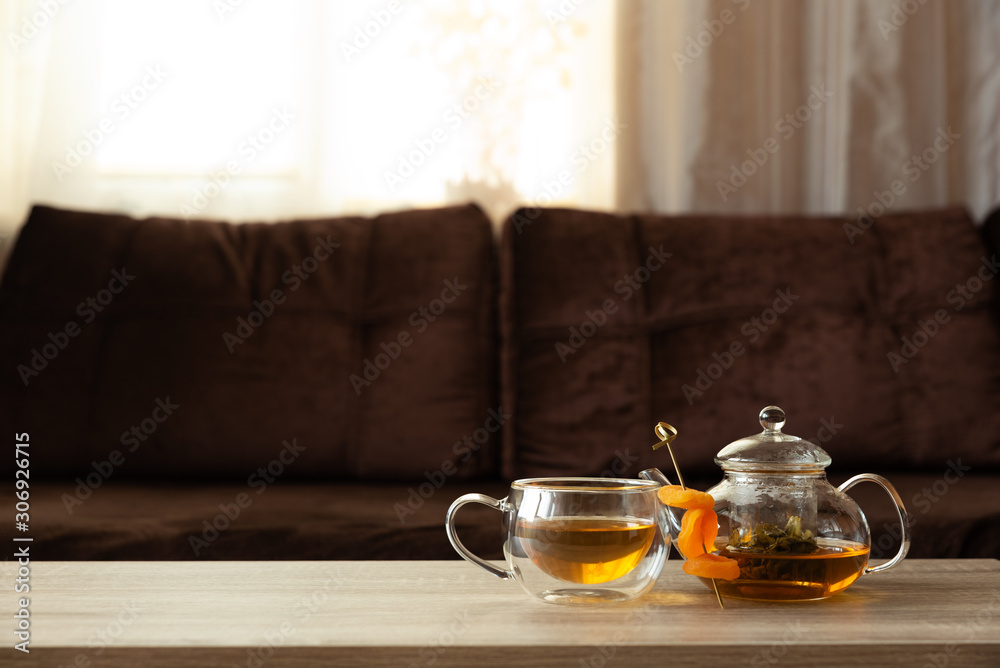 Glass teapot and thermo cup with green tea and dried apricots on wooden  table against defocused sofa with pillows. Front view. Mock-up Stock Photo