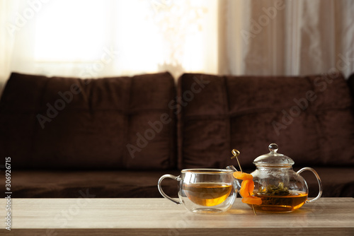 Glass teapot and thermo cup with green tea and dried apricots on wooden table against defocused sofa with pillows. Front view. Mock-up