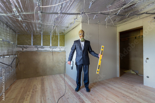 Strange man in businessman suit and gas protection mask inside a room under renovation works holding electric screwdriver and a level tools. photo