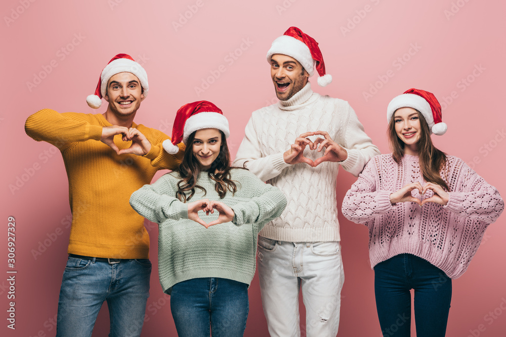 happy friends in sweaters and santa hats showing heart symbols, isolated on pink