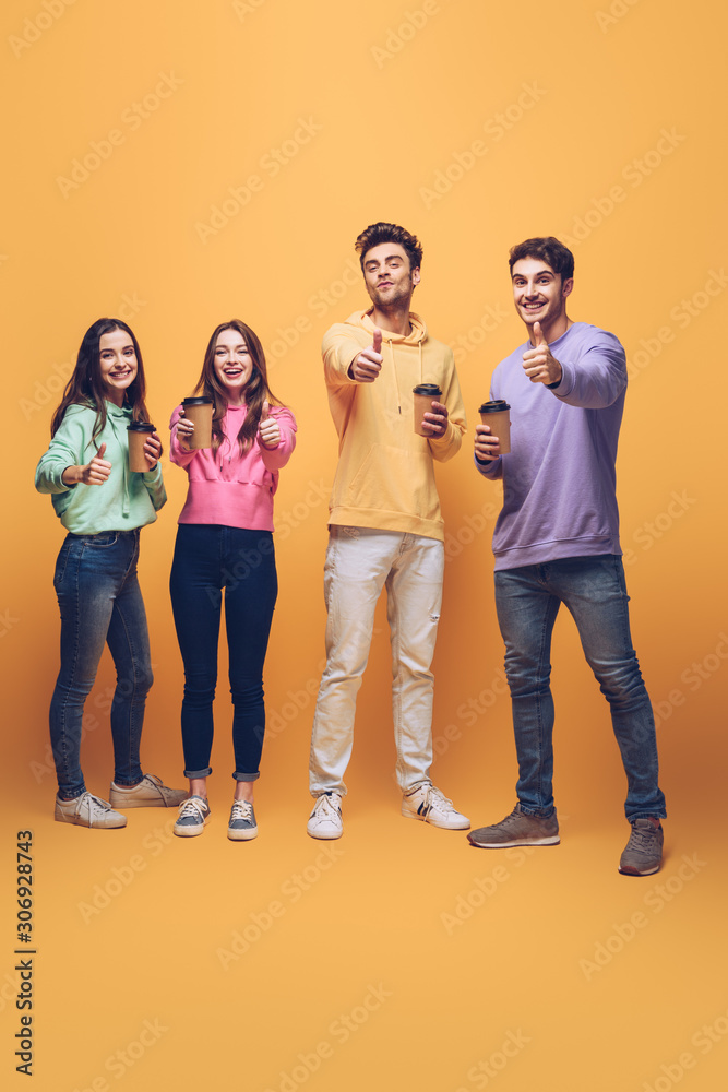 Fototapeta premium smiling friends holding coffee to go and showing thumbs up, on yellow