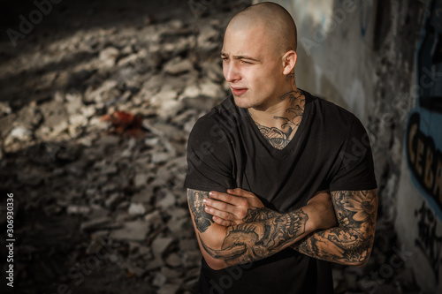 A young handsome bald brutal guy stands against the backdrop of the ruins of a building. Tattooed attractive man with arms fully covered by tattoos in a ruined building. Concept of crime, hooliganism.