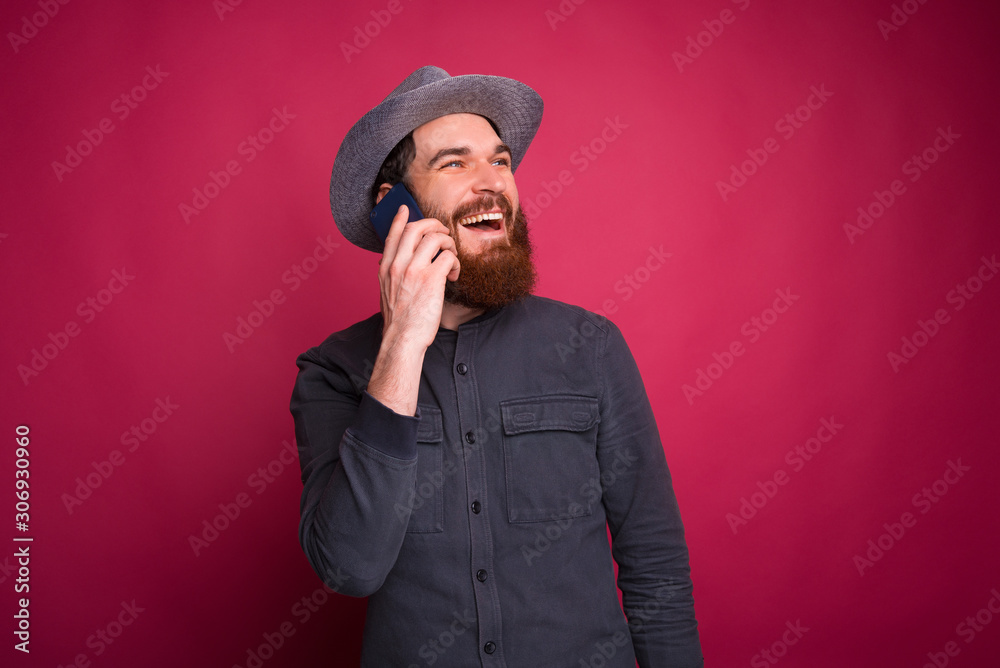 Young bearded man looking away, smiling and talking to the phone near pink wall. Young bearded man looking away, smiling and talking to the phone near pink wall.