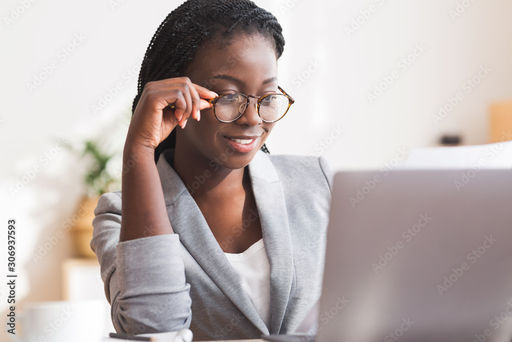 Afro secretary wearing glasses and working with papers and laptop - obrazy, fototapety, plakaty 
