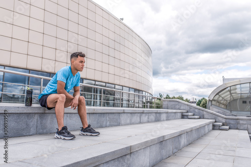 male athlete sits on steps and looks into distance, rests after workout and enjoys view, workout workout, in summer city. Fitness motivation youth lifestyle. Free space for copy text.