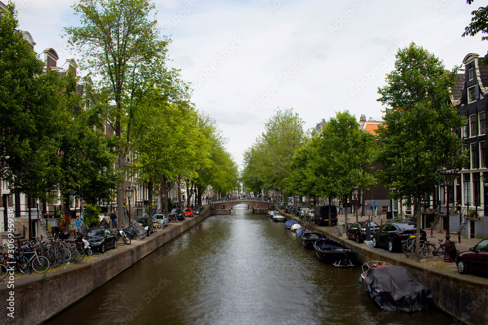 View of canal, parked boats, cars and bicycles, people walking on street, trees and historical, traditional buildings in Amsterdam. It is a sunny summer day.