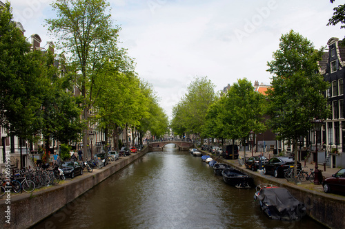 View of canal, parked boats, cars and bicycles, people walking on street, trees and historical, traditional buildings in Amsterdam. It is a sunny summer day. © theendup