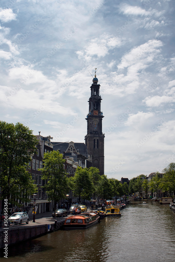 View of canal cruise tour boats, Westerkerk church's tower, trees and historical, traditional buildings. It is a sunny summer day.