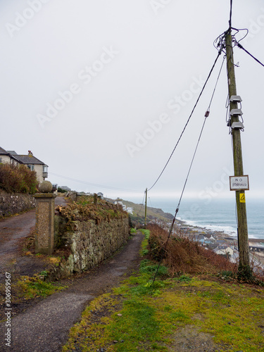 Wide vertical view of an old cliffside path overlooking the town of Sennen Cove on a cloudy day. Cornwall, United Kingdom. Travel and tourism.