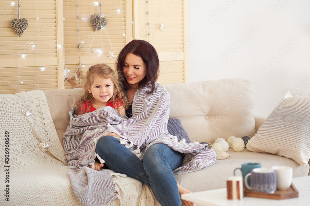 mother and daughter at home photo. Young mother sits on sofa with plaid and kisses her little daughter. Christmas garland and decorations.