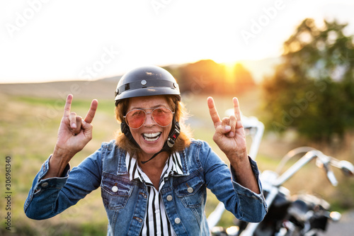 A cheerful senior woman traveller with motorbike in countryside at sunset.