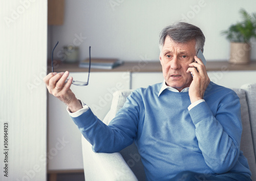 Senior Man Having Unpleasant Phone Conversation Sitting On Couch Indoor photo