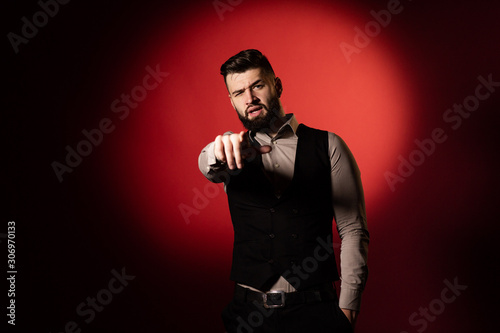 Portrait of a young serious bearded man in a black vest on red background. Man pointing at the camera