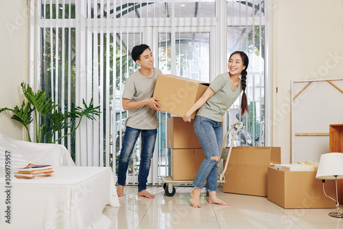 Happy young Vietnamese couple in casual clothes carrying heavy cardboard box when moving in new house