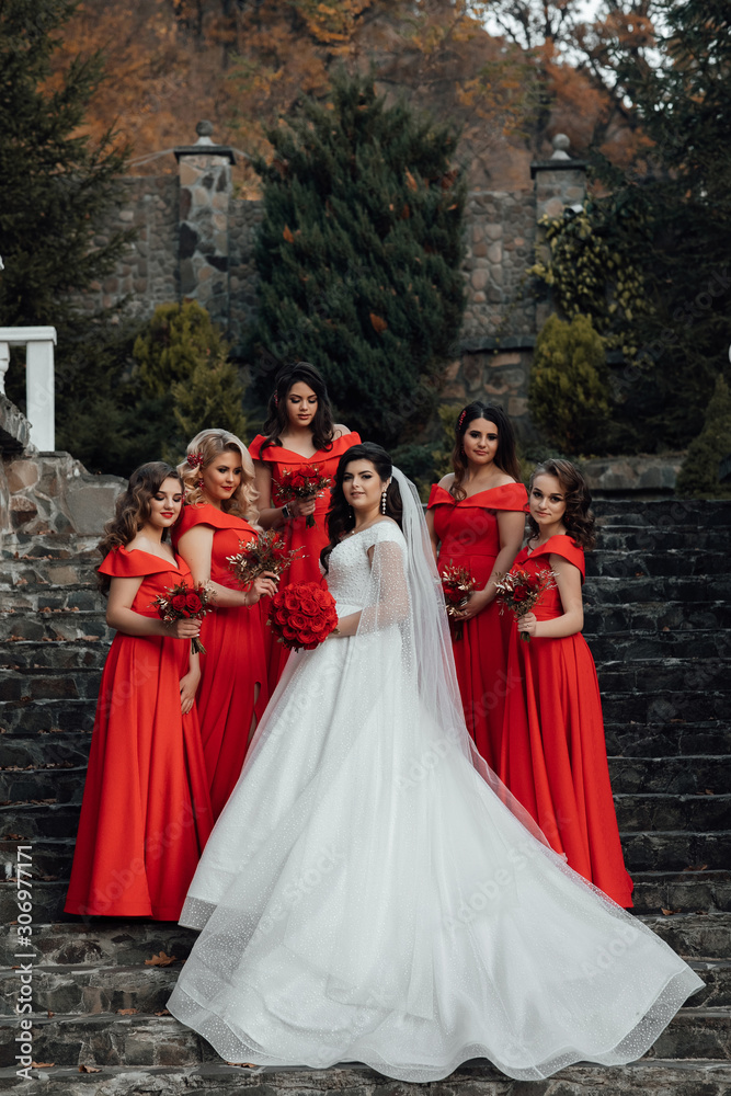 Group portrait of bride and bridesmaids on the stairs. Wedding in autumn. Bride in wedding dress and bridesmaids in red dresses at wedding day. Stylish wedding in red color. Marriage concept.
