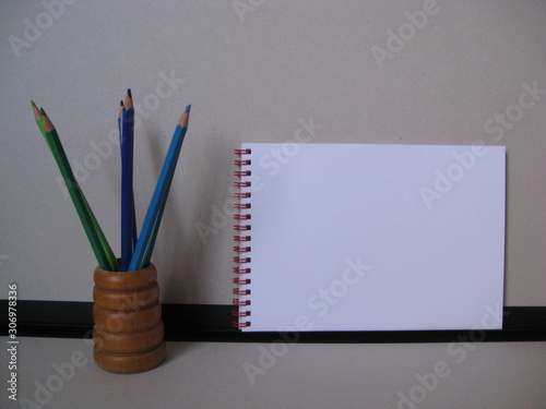 lápices de madera con minas de varios colores gama azules y verdes,  en porta lápices de madera marrón miel con ribetes circulares, con fondo gris, negro, y cuaderno de anillas rojas para dibujar  photo