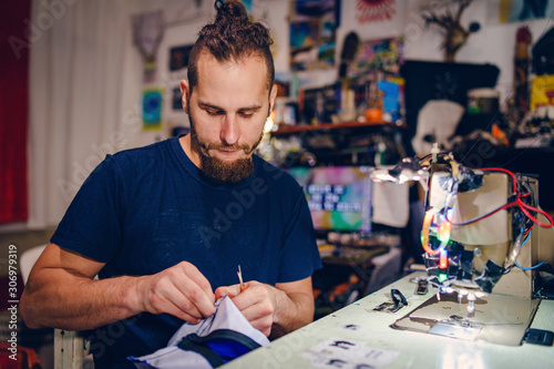 Adult man tailor with beard sitting by his sewing machine at his work shop making a bag checking stitches handmade creative craft working at home