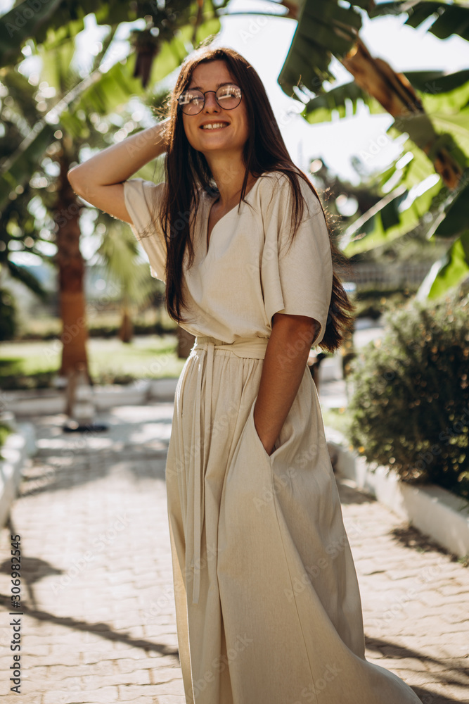 stylish young attractive woman dressed in a long linen dress with a hat photographed in the summer on a background of palm trees and the sea in the bright sun