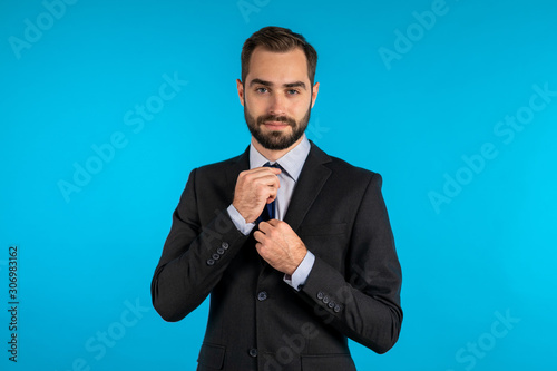 Handsome young businessman corrects his necktie and looking to camera like in mirror. Man on blue studio background. Corporate person in elegant suit correcting his tie.