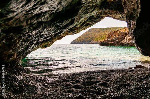 Amazing seascape view from unique sea cave overhanging above water on famous Cala Bianca beach scenic surroundings. Crystal clear sea water waves washing pebbles inside the massive rocky cave