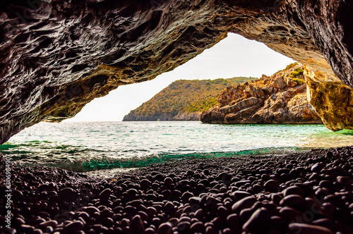 Amazing seascape view from unique sea cave overhanging above water on famous Cala Bianca beach scenic surroundings. Crystal clear sea water waves washing pebbles inside the massive rocky cave photo