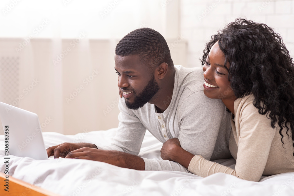 Cheerful afro spouses lying in bed using laptop together