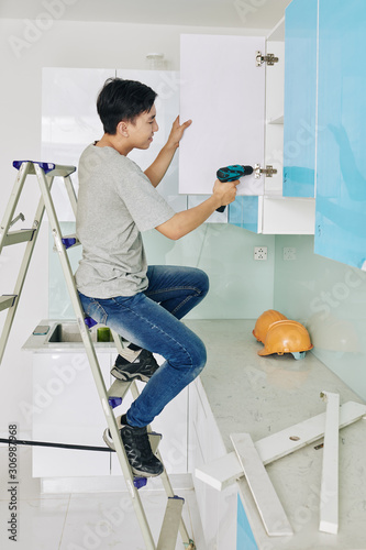 Young Asian man sitting on ladder when fixing door of kitchen cupboard