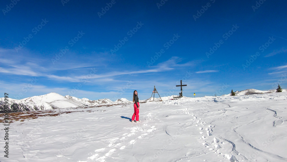 A girl in a skiing outfit going up a snowy hill towards the mountain's peak in Bad Kleinkirchheim, Austria. Snow caped Alps. The snow is gradually melting, giving a way to spring. Clear day.