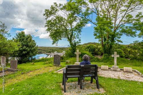 Mature woman sitting relaxed on a bench with her back to the camera, with graves, crypts, crosses in an ancient cemetery on a hill with Clifden bay in the background, sunny spring day in Ireland