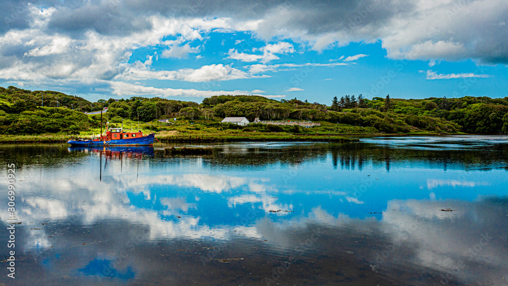 Obraz premium Clifden bay with clouds reflecting in the water and a boat anchored in the water at high tide, surrounded by green vegetation, spring day with a blue sky and white clouds Ireland