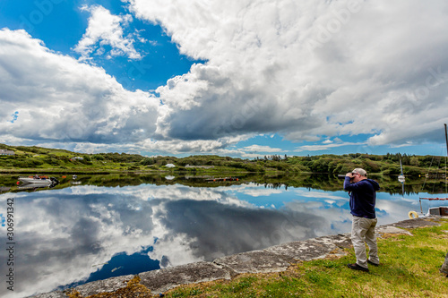 Photographer taking a picture in the port of Clifden at high tide, anchored ships and clouds reflecting in the water, sunny spring day with a blue sky and abundant white clouds and Clifden, Ireland