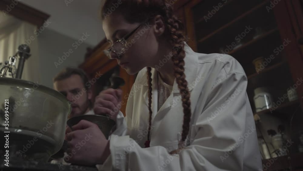 Bottom view of beautiful Caucasian girl with pigtails working in ancient pharmacy. Young redhead woman grinding ingredients using mortar and pestle. Her male colleague sitting at the background. Bottom view of beautiful Caucasian girl with pigtails working in ancient pharmacy. Young redhead woman grinding ingredients using mortar and pestle. Her male colleague sitting at the background.