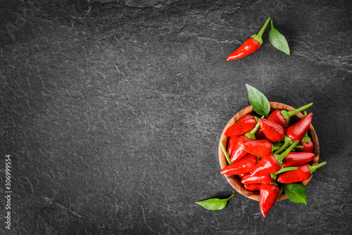 Red hot peppers in wooden bowl on dark stone table. Chili small spice pepper and green leaves on black background top view and copy space.