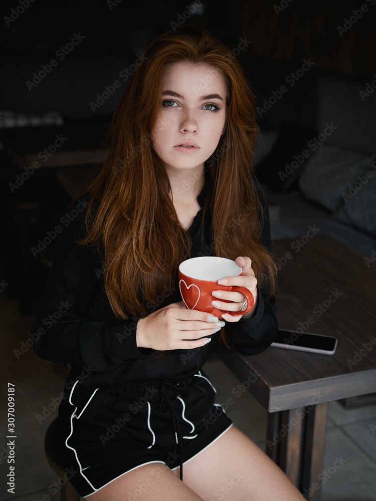 Cute redhead woman woman with adorable smile sitting in cozy city cafe enjoying free time coffee break holding big red mug in hands dressed black blouse and shorts. Cute redhead woman woman with adorable smile sitting in cozy city cafe enjoying free time coffee break holding big red mug in hands dressed black blouse and shorts.