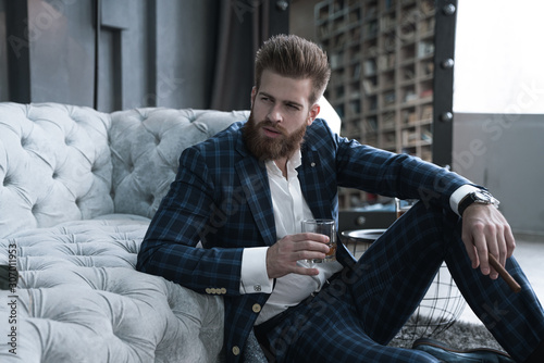 Handsome stylish man blue suit in a cage at home sitting on the floor with glass of whiskey in hand.