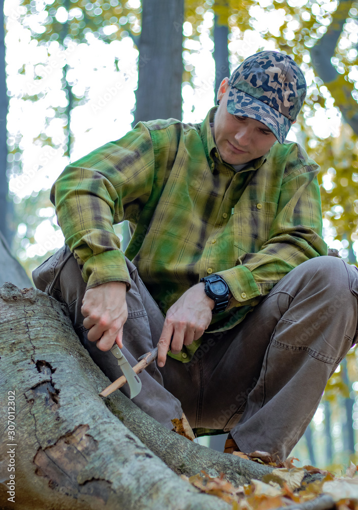Man with knife sharpening a wooden stick for campfire in the forest. Bushcraft, hunting and survival concept.