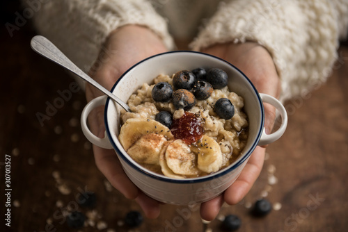 Woman hands offering a vegan porridge with oat flakes, banana, blueberries, chia, cinnamon, maple syrup and strawberry jam. Horizontal. Top view. photo