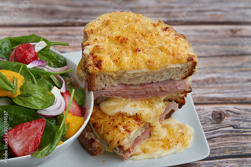 Sandwich with Parmesan and Gruyere cheese, ham, béchamel sauce and artisan bread accompanied by a salad on a wooden background.  The Croque Monsieur.