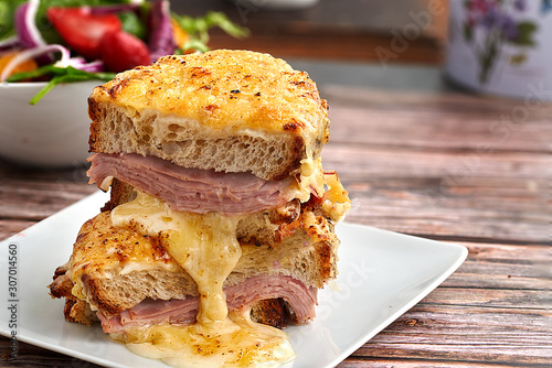 Sandwich with Parmesan and Gruyere cheese, ham, béchamel sauce and artisan bread accompanied by a salad on a wooden background.  The Croque Monsieur.