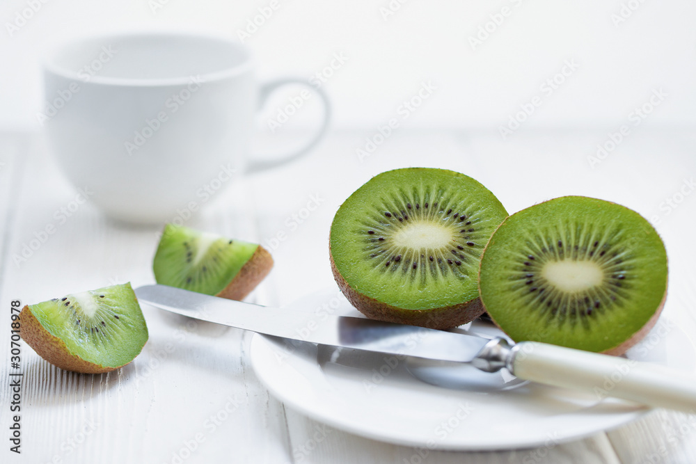 Sliced fresh kiwi fruit on a white plate on a light wooden table with a knife and white cup on background. Front view. Sliced fresh kiwi fruit on a white plate on a light wooden table with a knife and white cup on background. Front view.