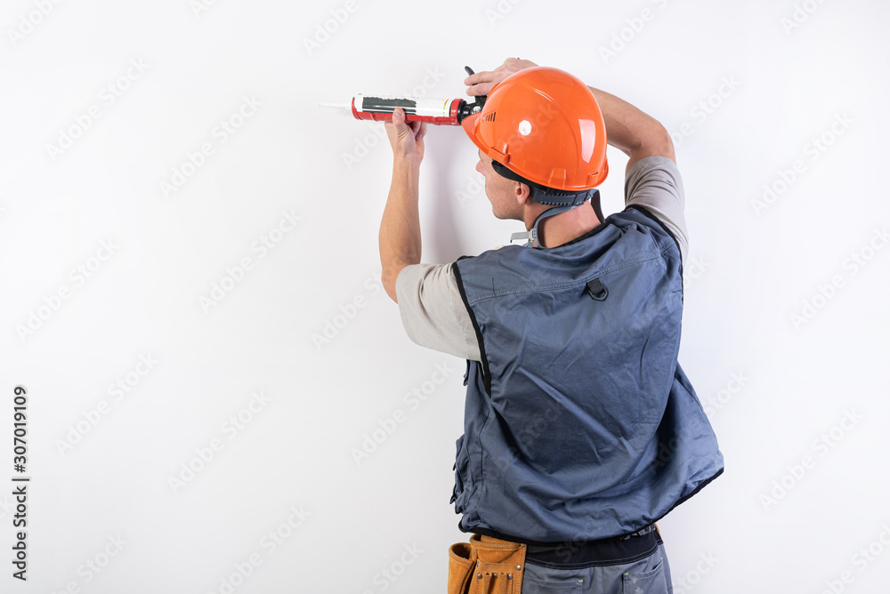 A builder in a helmet, with silicone in a mounting gun, puts on the wall. On a light background.