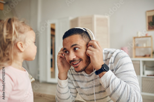Portrait of young dad showing big headphones to cute little girl while playing together at home, copy space