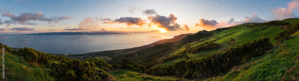 Panoramic view of an Azores sunset across the hilly São Jorge countryside with Pico Island across the sea in the background with the summit of Mount Pico visible on the horizon within dramatic clouds.