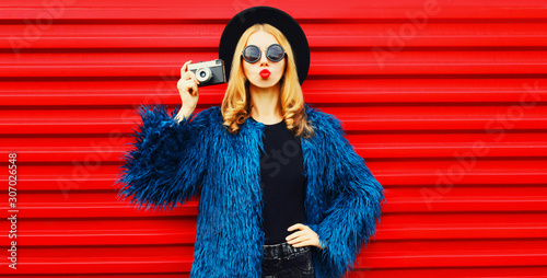 Portrait stylish woman with retro camera blowing red lips sending sweet air kiss wearing blue faux fur coat, round hat and sunglasses over colorful red wall background © rohappy