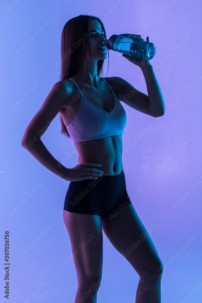 Young sporty muscular woman drinking water, isolated against purple light background Young sporty muscular woman drinking water, isolated against purple light background