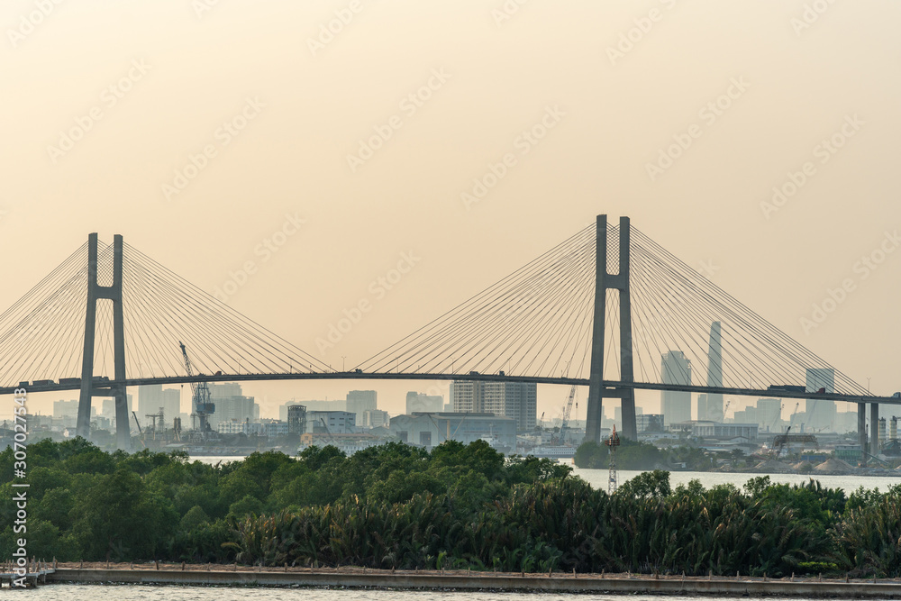 Ho Chi Minh City, Vietnam - March 13, 2019: Sunset sky shot over Song Sai Gon river. Phu My suspension bridge with downtown skyscraper buildings on horizon, and green foliage in front..
