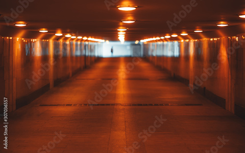 View with a shallow depth of field of a long underground pass with three rows of lamps stretching into the vanishing point with a human silhouette at the end, selective focus in the foreground