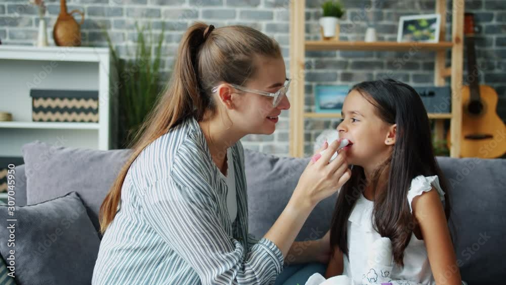 Loving mother is putting lip gloss on little daughter's lips smiling talking at home expressing love and care while small girl is enjoying women's activity. Beauty and childhood concept. Loving mother is putting lip gloss on little daughter's lips smiling talking at home expressing love and care while small girl is enjoying women's activity. Beauty and childhood concept.
